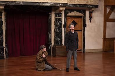 ASC_25_King Lear_012-Fool_Lear Lear, wearing black shirt and gray pants, stands stalwart with head raised while the Fool in houndstooth cap and wrapping Lear's brown overcoat around herself kneels on the otherwise bare stage.